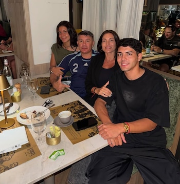 Cuatro personas, dos hombres y dos mujeres, sentadas en una mesa de restaurante, sonriendo a la cámara. Hay platos con comida, copas y teléfonos