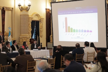 Sala formal con columnas y candelabros. Un gráfico económico se proyecta en pantalla. Varias personas sentadas observan la presentación en la reunión