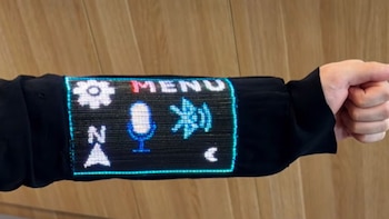 Brazo con una manga negra que contiene una pantalla luminosa con la palabra MENU y diversos iconos digitales, con el puño cerrado