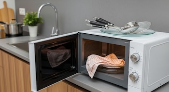 Un microondas blanco con la puerta abierta contiene un trapo de cocina. Sobre el microondas hay un plato de vidrio con utensilios de metal. Al fondo, una cocina moderna.