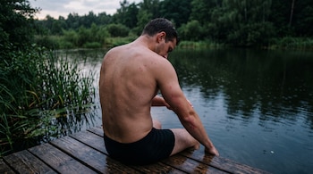 Vista posterior de un hombre sin camiseta sentado en un muelle de madera, mirando la irritación roja en su antebrazo junto al agua.