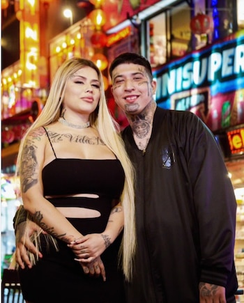 Mujer rubia con vestido negro y hombre con chaqueta negra, ambos con tatuajes visibles en cara y brazos, posan sonriendo frente a un fondo urbano iluminado