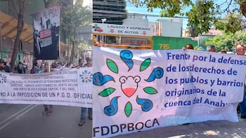 Manifestantes sostienen pancartas en una calle bajo un cielo azul claro, con edificios y árboles al fondo. Las pancartas tienen texto y un logo