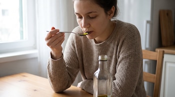 Una mujer joven con suéter beige toma una cucharada de líquido amarillo. Una botella de vidrio con el mismo líquido está sobre una mesa de madera.
