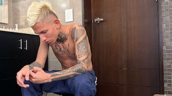 Hombre joven, sin camisa, con cabello rubio y tatuajes, sentado, con pantalones azules, en una habitación con puerta de madera, lavabo y espejo.