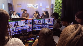 Personas en un estudio de televisión. Múltiples monitores LCD muestran una transmisión en vivo con presentadores, micrófonos, audífonos y equipos de producción