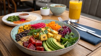 Buddha bowl colorido con tofu, aguacate, remolacha, frijoles negros, quinoa, maíz y vegetales sobre mesa de madera. Zumo de naranja y otros platos al fondo.