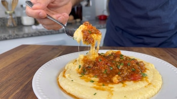 Una mano sostiene un tenedor levantando polenta cremosa con salsa de carne y perejil picado, con hebras de queso, de un plato blanco.