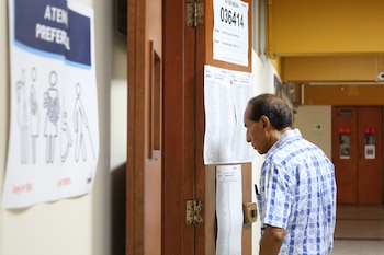 Un hombre de camisa a cuadros azul y blanca mira una lista de electores pegada en una puerta de madera marrón con bisagras metálicas