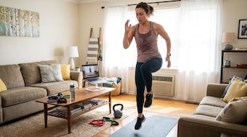 Mujer en ropa deportiva saltando sobre una colchoneta, computadora portátil, botella de agua, bandas elásticas y pesa rusa en una sala de estar.