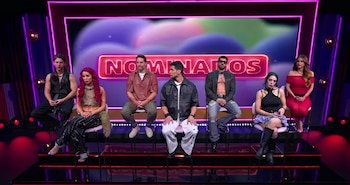 Siete personas, cinco hombres y dos mujeres, sentadas en taburetes sobre un escenario iluminado con luces de neón. Detrás, una pantalla muestra la palabra NOMINADOS en rojo