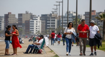 Varias personas pasean y se relajan en una playa soleada con un malecón. Edificios urbanos y palmeras se ven al fondo.