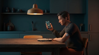 Hombre sentado en una mesa, mira su smartphone con gráfico de glucosa. Un sensor de monitoreo está en su brazo. Un plato vacío y cubiertos sobre la mesa.
