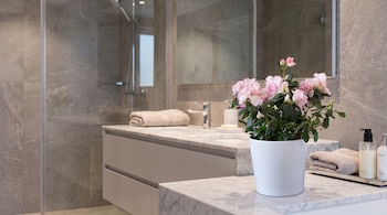Baño moderno con paredes de azulejos grises, mampara de ducha de cristal, lavabo doble con encimera de mármol, espejo, toallas y una maceta con flores rosadas.