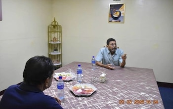 Dos hombres se abrazan de espaldas a la cámara junto a una puerta abierta en una habitación con paredes claras, una mesa y estantería