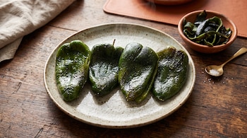 Cuatro chiles poblanos asados en un plato de cerámica, un cuenco de barro con tiras de piel y una cuchara con sal sobre una mesa de madera.