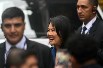 Keiko Fujimori sonríe, con dos hombres de traje y audífonos visibles detrás. Hay personas borrosas en el primer plano y fondo