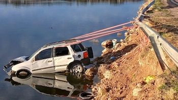 Un vehículo utilitario deportivo blanco semihundido en el agua. Se observan cuerdas rojas de rescate, un guardarraíl metálico dañado y tierra al costado