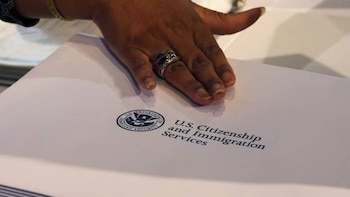 Primer plano de una mano con un anillo sobre un documento blanco que muestra el sello del Departamento de Seguridad Nacional y las palabras USCIS