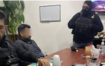 Un hombre con el rostro difuminado permanece sentado junto a una mesa frente a un agente con el rostro cubierto y chaleco táctico