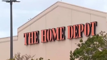 Primer plano de la fachada de una tienda The Home Depot con su icónico letrero naranja y negro, un poste de luz a la izquierda y follaje verde en la parte inferior