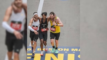 Tres corredores masculinos cruzan la línea de meta de la Maratón de Boston. El corredor central es apoyado por los otros dos, con el logo "BOSTON MARATHON" visible en el suelo