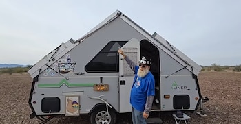 Un hombre con barba blanca y gafas de sol señala una caravana gris de techo triangular sobre un suelo pedregoso