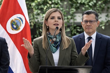 Mujer con blazer verde y bufanda, hablando frente a un micrófono. Bandera de Costa Rica a la izquierda y un hombre con gafas en el fondo.