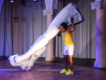 Un hombre en camiseta amarilla y shorts blancos carga una enorme escultura de pierna sobre sus hombros en un escenario con columnas y cortinas