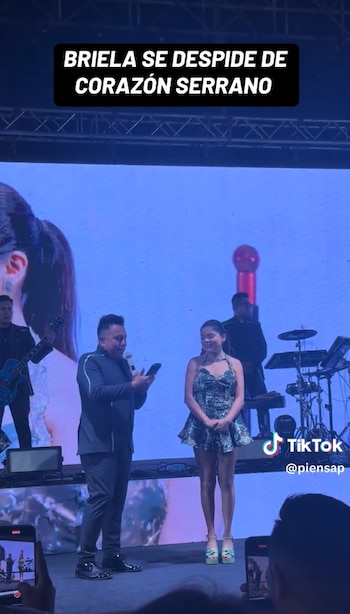 Una cantante con vestido corto estampado y un hombre con micrófono están sobre un escenario frente al público