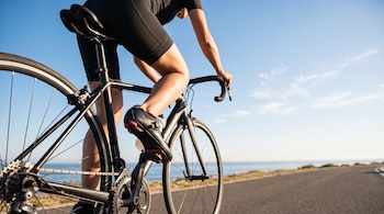 Plano bajo de una persona en una bicicleta de carreras negra pedaleando por una carretera asfaltada junto al mar.