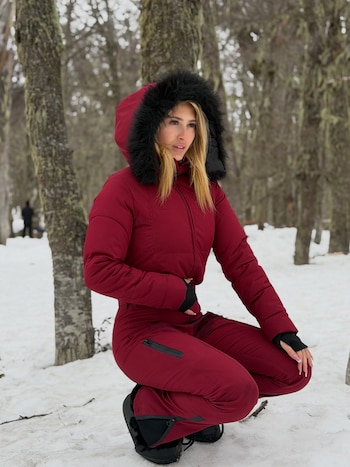 Una mujer posa de cuclillas sobre la nieve entre árboles, vistiendo un traje térmico rojo con capucha con piel negra
