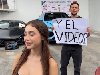 Una mujer con cabello largo y un hombre posan. Él sujeta un cartel con "Y EL VIDEO?". Detrás, dos coches deportivos y un letrero de "ELIMINATORIAS MUNDIAL 2026"