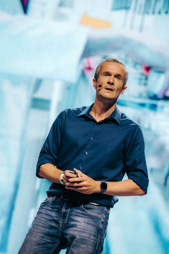 Hombre de mediana edad con camisa azul y jeans, cabello gris, micrófono en mejilla, reloj inteligente y pulsera. Sostiene un objeto oscuro con ambas manos