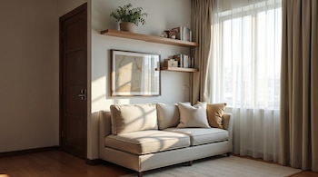 Sala pequeña y luminosa con sofá beige de dos plazas, estantes flotantes con libros y planta, puerta de madera oscura y ventana grande con cortinas claras.