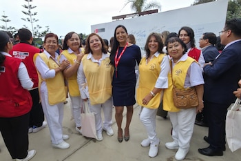 Un grupo de ocho mujeres sonríe a la cámara, de pie al aire libre. Una mujer central en vestido azul oscuro, y otras siete en chalecos amarillos sobre ropa blanca. Detrás hay una pared blanca con grafitis y árboles