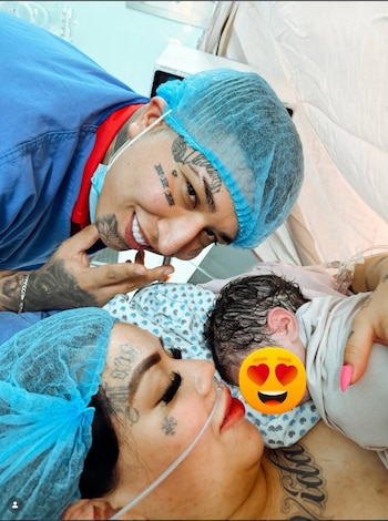 Hombre sonriente y mujer con cánula de oxígeno y gorros quirúrgicos miran a un bebé recién nacido. La mujer sostiene al infant en su pecho, ambos tienen tatuajes