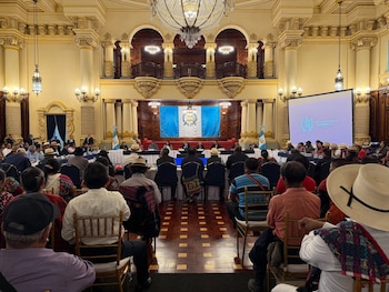 Público sentado de espaldas a la cámara en un salón. Al frente, una mesa con personas, banderas de Guatemala y una pantalla con el logo del gobierno