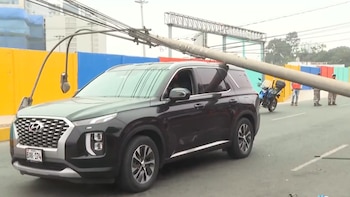 Un SUV negro, placa BXK-174, con un poste de alumbrado público caído sobre su parte delantera y el techo. Al fondo, una motocicleta azul y dos personas de pie