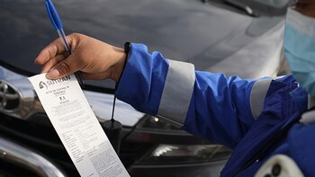 Una mano sostiene un bolígrafo azul y un acta de control de Sutran con el código F.1, mientras una persona con chaqueta azul con bandas reflectantes fiscaliza un vehículo