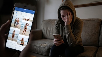 Mano sosteniendo un móvil con imágenes de cuerpos delgados en redes sociales. Al fondo, una persona con capucha se tapa la cara angustiada en un sofá.