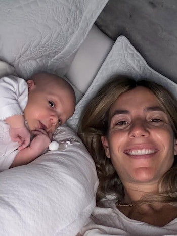 María Belén Ludueña sonríe a la cámara mientras yace junto a su bebé, Vito, acostado sobre sábanas blancas con un mameluco claro