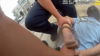 Hombre (Zach Murphy) esposado por un agente de policía en la calle. Primer plano de manos, esposas y uniforme. Al fondo, edificios y datos de cámara corporal