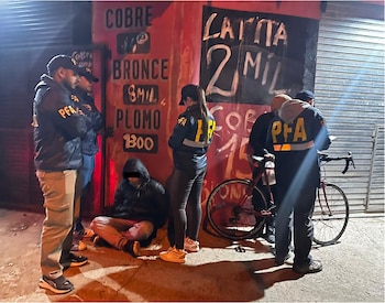 Cuatro agentes de PFA rodean a una persona sentada en el suelo, frente a un edificio con carteles de precios de metales y una bicicleta