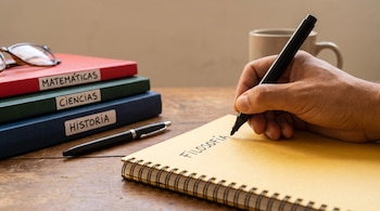 Una mano adulta escribe 'Filosofía' en un cuaderno amarillo con marcador negro. Hay tres cuadernos apilados, gafas, un bolígrafo y una taza.