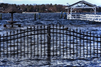 Una valla de metal negro está parcialmente sumergida en el agua. Al fondo, se ven un muelle blanco y una sombrilla de paja sobre el agua