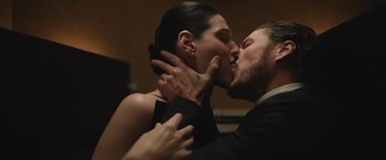 Un hombre y una mujer de perfil besándose en un primer plano. El hombre usa traje, la mujer vestido oscuro. Las manos del hombre sujetan el cuello de la mujer
