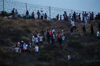 Múltiples hombres jóvenes de pie en una ladera con vegetación seca, frente a una valla de alambre de espino bajo un cielo nublado al anochecer
