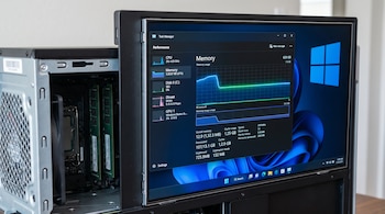 Computadora de escritorio con gabinete abierto y módulos RAM visibles. El monitor muestra el Administrador de tareas de Windows 11 en la sección de rendimiento de memoria.
