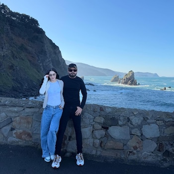 Un hombre y una mujer posan de pie junto a un muro de piedra con el mar, acantilados y formaciones rocosas al fondo bajo un cielo azul